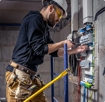 Electrician wearing yellow safety goggles and a white hard hat, working on an electrical panel with exposed wires.